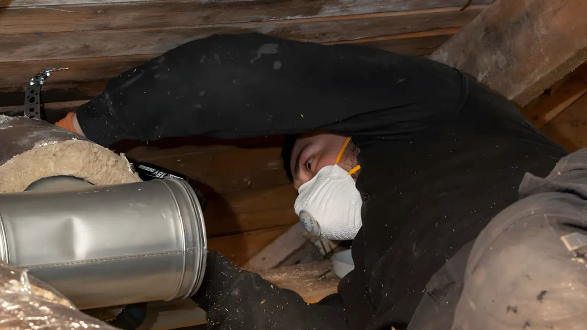 Tekniker med andningsmask ligger i ett trångt vindsutrymme i Stockholm och arbetar med en isolerad ventilationskanal